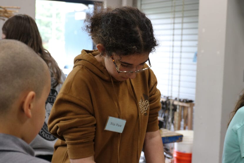 A student in a brown sweatshirt and glasses looks down in a classroom. Her sweatshirt has a nametag that says "Nia Paw."