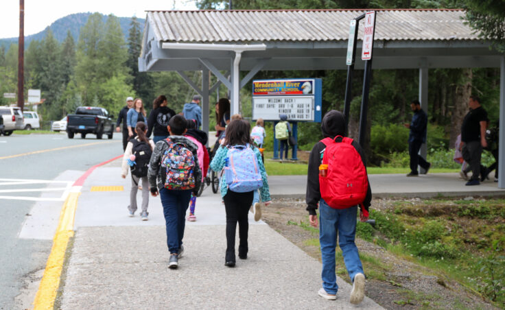 Several students walk on a sidewalk toward a covered walkway while carrying backpacks.