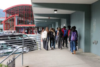 The backs and bags of a group of students walking under a covered sidewalk toward a school made of red metal and glass.