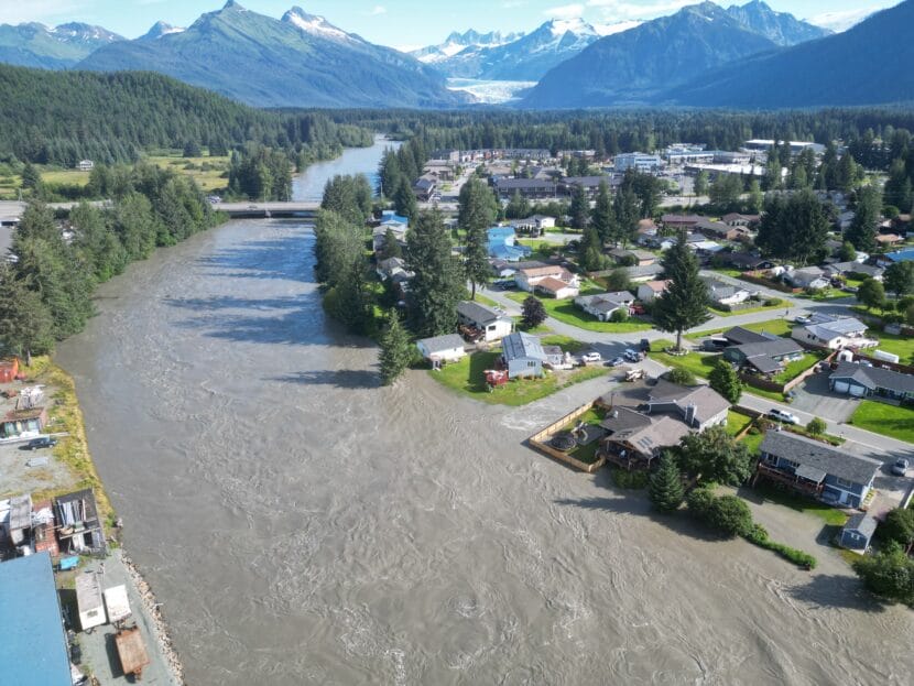Water flows down a river with homes on either side, showing some overflow onto surrounding streets. a glacier sits miles away in the background
