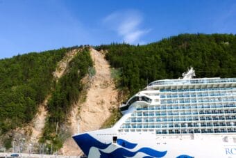 A large cruise ship in the foreground, with a steep hillside gashed with bare rockslide paths right behind it.
