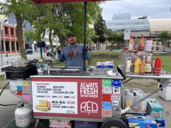 man poses in front of a hotdog stand on an urban street.