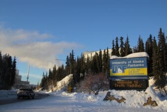 A winding road leading up to a large building with a large smokestack. A sign that says "University of alaska Fairbanks" is in the foreground with "Welcome to Troth Teddha'" on a light-up sign. The scene is snowy with spruce trees on a hillside