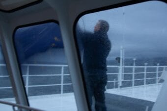 A man on a ferry deck, seen through a rain-splattered window, brings down an Alaska state flag in the rain.