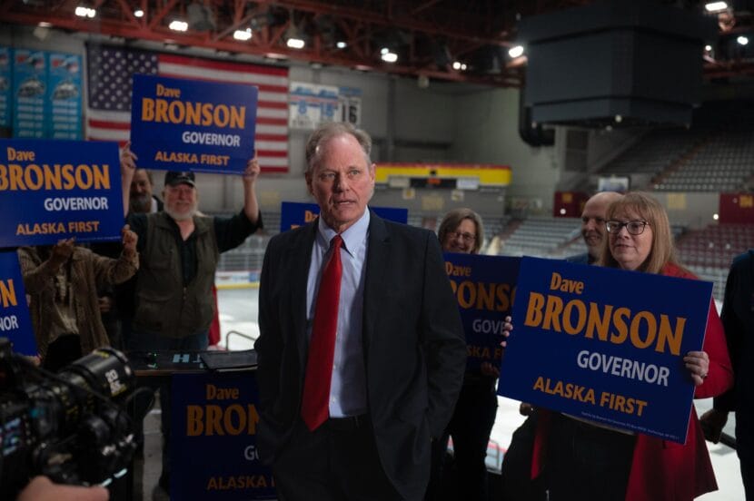 A man in a suit speaking in a hockey stadium surrounded by people holding blue signs with text that says "Dave Bronson, Governor, Alaska First"