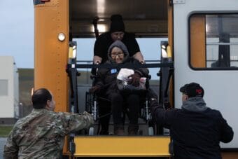 People in uniform help a woman in a wheelchair down from a bus ramp