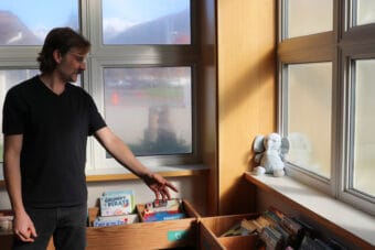 A man in a black T-shirt extends his arm toward picture books on short, wooden shelves.