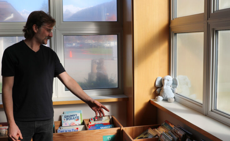 A man in a black T-shirt extends his arm toward picture books on short, wooden shelves.