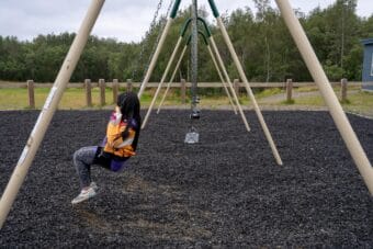 A girl in a yellow jacket with long black hair is the only child swinging on an outdoor swing set.