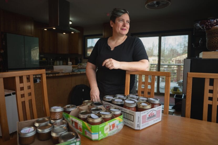 A woman in a black shirt poses with home made jam in her kitchen. 