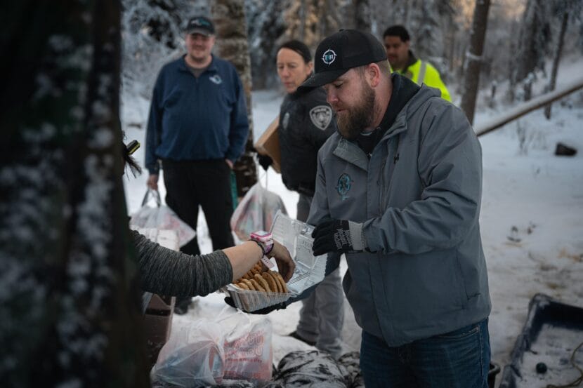 A man in a grey jacket offers a plastic box of cookies to a person out of frame. 