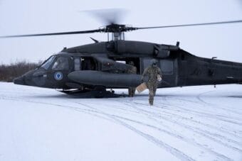 A person wearing camo and carrying a cardboard box walks away from a military helicopter in a snow-covered flat area