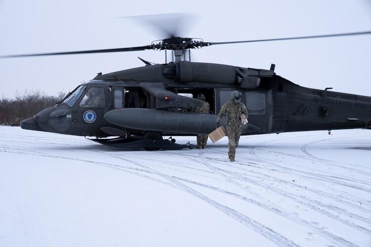 A person wearing camo and carrying a cardboard box walks away from a military helicopter in a snow-covered flat area
