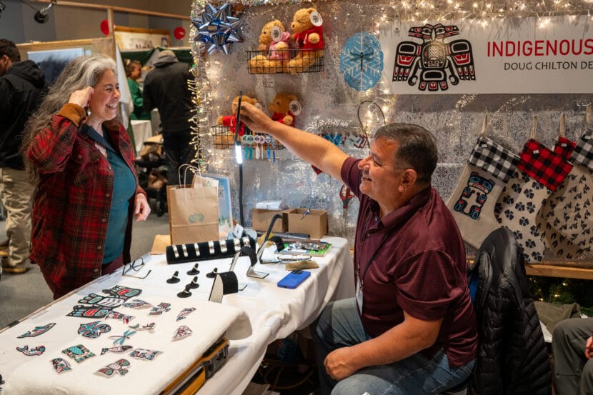 Doug Chilton holds up a mirror for a customer to see a pair of silver earrings at Juneau Public Market on November 29, 2025. (Photo by Alix Soliman/KTOO)