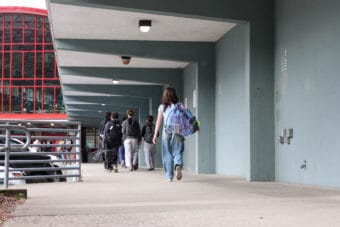 A student in jeans carrying a large purple and blue backpack walks on a covered walkway toward the front entrance of Juneau-Douglas High School: Yadaa.at Kalé.