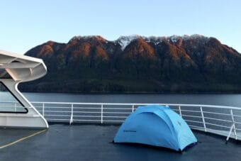 A small tent pitched on the deck of a ferry, with coastal mountains in the background.