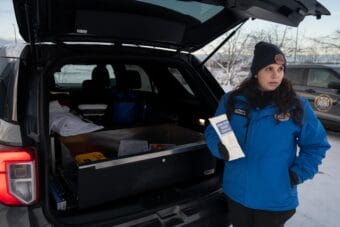 A woman holds up an opioid overdose kit next to a grey SUV with the trunk open.