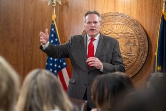 Man in wood-paneled room wearing a suit standing in front of state seal and flags
