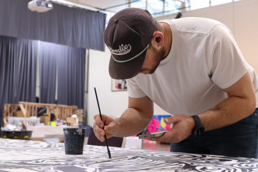 Rio Schmidt, dressed in a baseball cap, fills in a large block print mural with black paint.