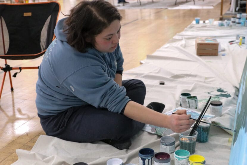 A person dressed in a blue sweatshirt sits cross-legged on the floor and paints a mural.