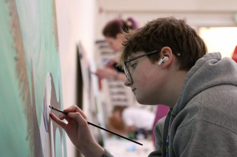 A teenager wearing glasses and a gray sweatshirt paints a large orca.
