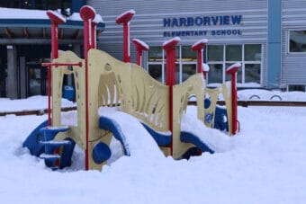 A red, blue and yellow play structured is buried in snow with Harborview Elementary School in the background.