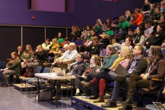 People sit in raised auditorium seats in front of a purple wall.