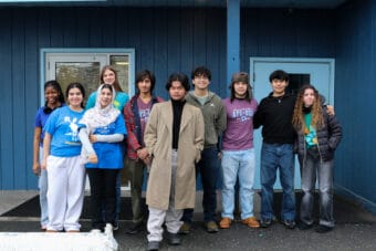 Ten students stand together outside of a blue building.