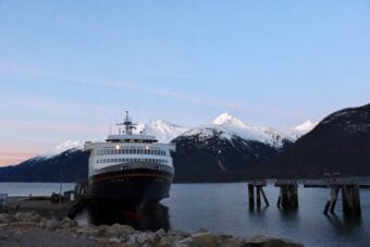 A docked Alaska state ferry with snowy mountains in the background, across a body of water.
