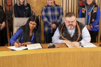Aparna Palmer and Chaylee Éesh Richard Peterson sign separate copies of a memorandum of understanding with several university staff standing behind them.