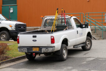 A white pickup truck is parked outside a brown building. It is filled with various maintenance and landscaping tools.