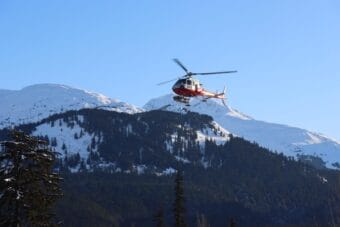 A red-and-white helicopter with a ski basket descending to land with snowy mountains in the background