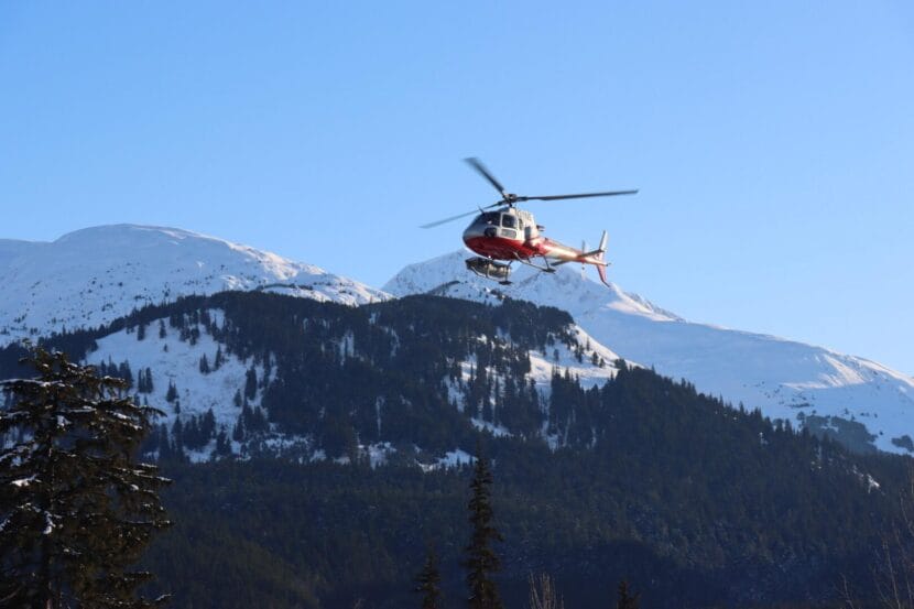 A red-and-white helicopter with a ski basket descending to land with snowy mountains in the background