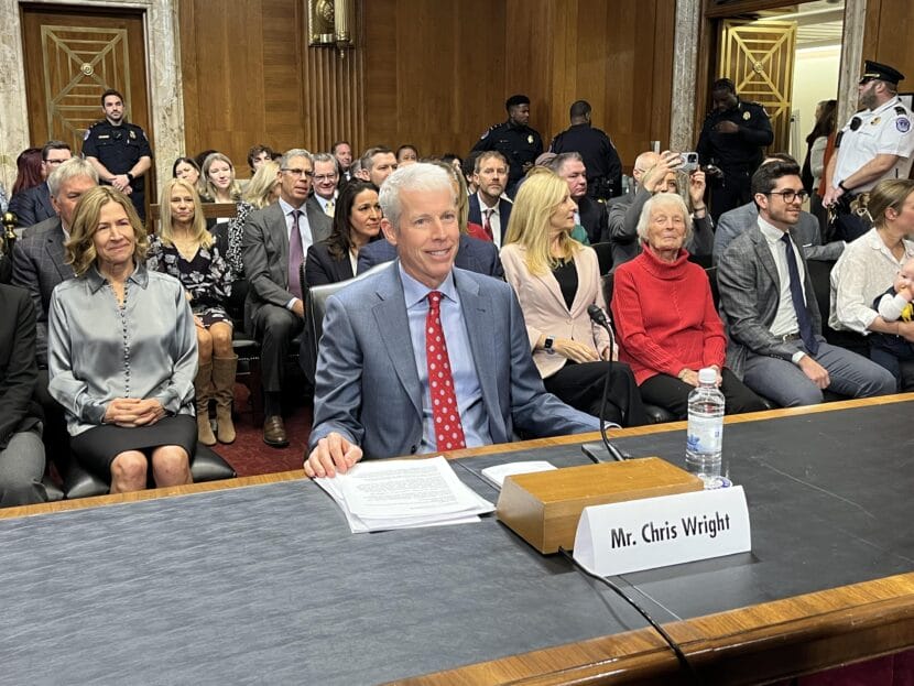 Man in a suit at a table, with lots of people seated behind him.