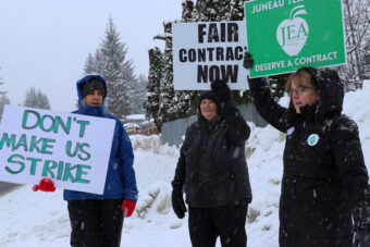 Three people dressed in dark jackets stand in the stay and hold up signs that say, "Don't make us strike," "Fair contract now," and "Juneau teachers deserve a contract."