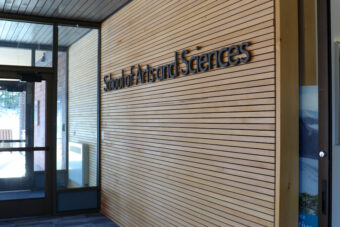 A sign in black lettering says "School of Arts and Sciences" on a wood paneling wall.