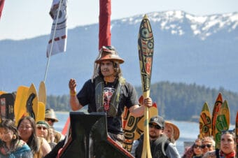 A young man with a painted, carved Raven hat and a formline painted paddle speaks from the bow of a yaakw or canoe. Ḵaayák’w Brandon Gomez introduces the Wind Dancer yaakw and asks permission to come ashore at Auke Recreation Area on June 2, 2026. (Photo by Yvonne Krumrey/KTOO)