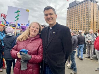 man in clerical collar and a woman in a raspberry-red jacket smile at camera