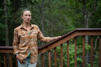 A woman wearing plaid stands, with a wooden bannister to her back, and gazes at the camera.