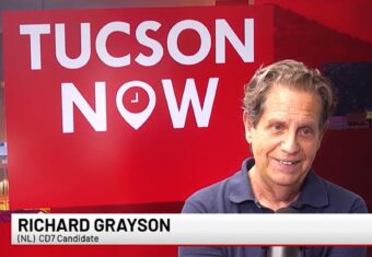 Richard Grayson is identified by a banner at the bottom of the image. Behind him is a red backdrop that says Tucson Now.